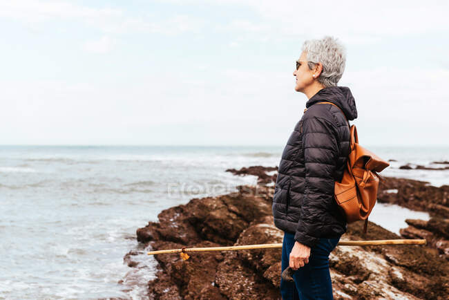 Seitenansicht einer lächelnden älteren Wanderin mit Sonnenbrille und grauen Haaren, die gegen stürmischen Ozean wegschaut — Stockfoto