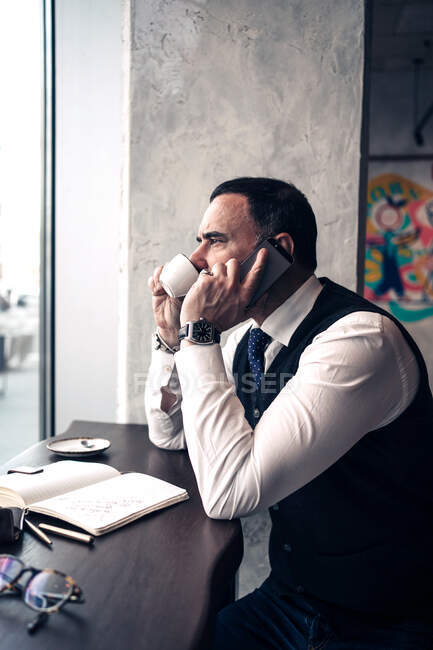 Side view of mature ethnic male entrepreneur talking on cellphone while drinking hot beverage and looking forward in cafeteria — Stock Photo