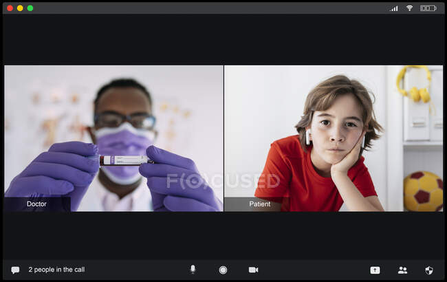 Концентрований молодий чорний лікар у медичній формі та маска, що показує пробірку з аналізом крові уважному хлопчику з вухами під час відеозв'язку — стокове фото