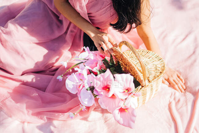 From above crop peaceful Asian woman in pink dress sitting on plaid with blooming flowers in wicker basket in lush garden and looking down — Stock Photo