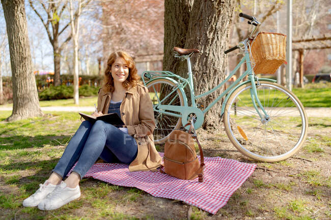 Ganzkörper glückliche junge Frau macht sich Notizen in Notizbuch auf kariertem Stoff mit Rucksack in der Nähe von Fahrrad im Park — Stockfoto