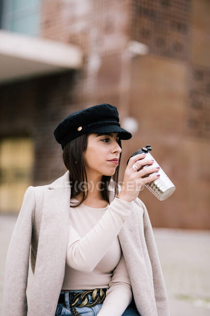 Female entrepreneur in trendy outfit sitting on bench with cup of beverage to go and looking away — Stock Photo