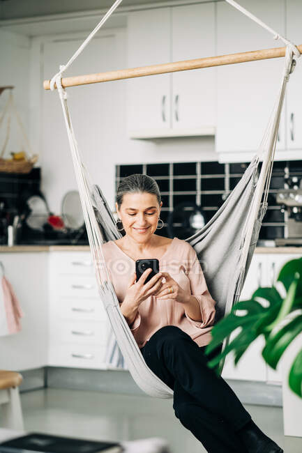 Positive Frau mittleren Alters surft Handy, während sie in einer bequemen Hängematte in der Nähe der Küchentheke mit Geschirr zu Hause mit grüner Pflanze sitzt — Stockfoto
