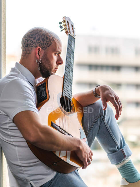 Hombre pensativo con la cabeza calva tatuada en ropa casual sentado en el alféizar de la ventana y tocando la guitarra en el día - foto de stock