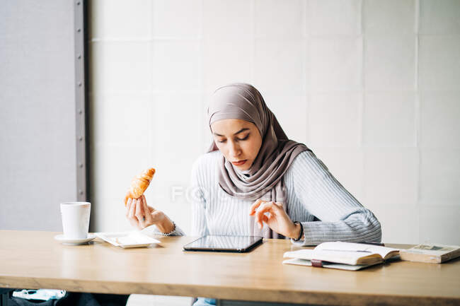 Ethnic female in hijab surfing Internet on tablet while sitting at table with croissant and cup of coffee in cafe and enjoying weekend — Stock Photo