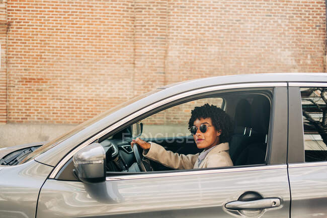 Vista lateral del conductor femenino afroamericano con gafas de sol elegantes y atuendo de moda sonriendo mientras conduce un automóvil moderno en la calle - foto de stock