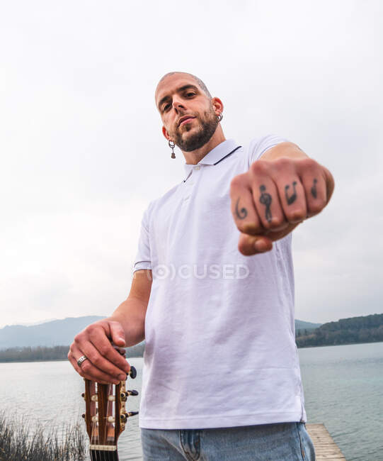 From below of bearded male musician showing tattoos on finders while standing with guitar on wooden pier and demonstrating fist in nature against lake and mountains in cloudy day — Stock Photo