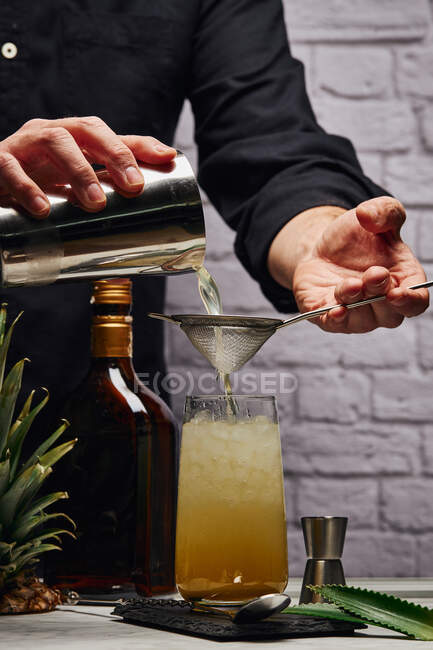 Cultivar barman irreconhecível derramando coquetel através de coador de coqueteleira em vidro cheio de cubos de gelo contra a parede de tijolo — Fotografia de Stock