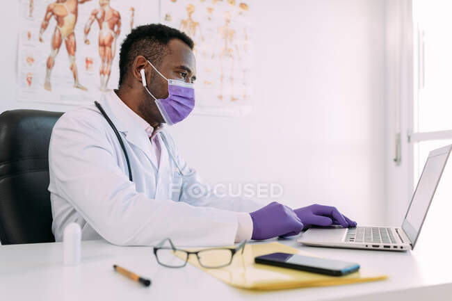 Médico masculino afro-americano concentrado em roupão médico e máscara trabalhando no laptop na clínica moderna — Fotografia de Stock