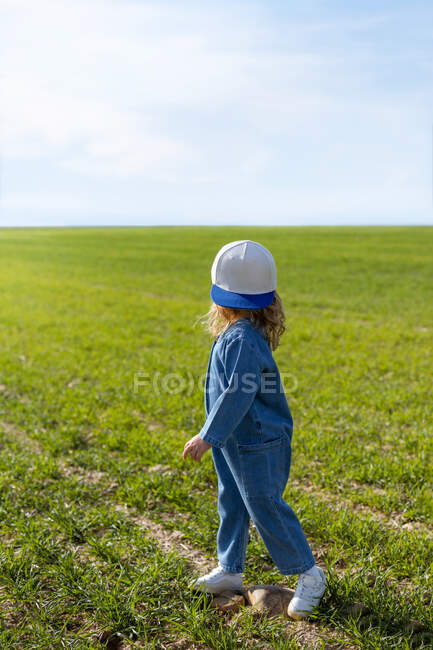 Ganzkörper-Seitenansicht des nicht wiederzuerkennenden Mädchens in stilvoller Kleidung und Mütze, die wegschaut, während sie an einem sonnigen Sommertag im Feld auf dem Gras steht — Stockfoto