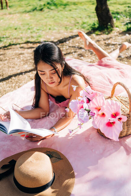 Ethnique asiatique femelle en robe élégante couché sur la couverture avec des fleurs dans le panier en osier et des chaussures avec chapeau de paille et livre de lecture dans le jardin — Photo de stock