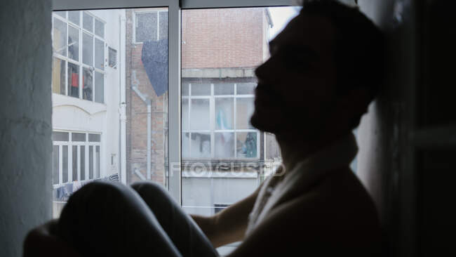 Боковой вид задумчивого мужчины, обнимающего колени и сидящего дома на подоконнике — стоковое фото