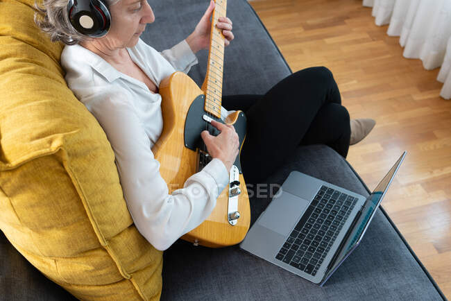 Vue arrière grand angle du guitariste féminin dans les écouteurs jouant de l'instrument de musique sur le canapé avec netbook à la maison — Photo de stock