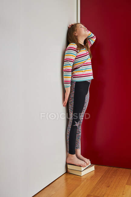 Side view of cheerful cute girl standing on a pile of books near wall and measuring her height and looking up — стоковое фото