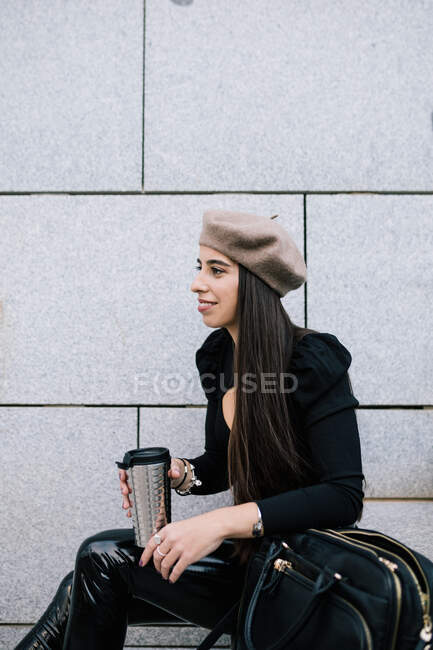 Vue latérale de ravie femme entrepreneur élégant en tenue à la mode et chapeau de béret assis sur les escaliers avec boisson pour aller en ville et détourner les yeux — Photo de stock