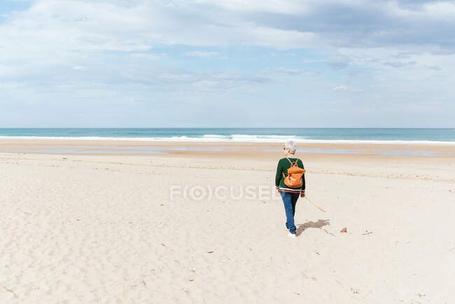 Back view of unrecognizable senior female trekker walking on sandy shore against endless sea during trip — Stock Photo