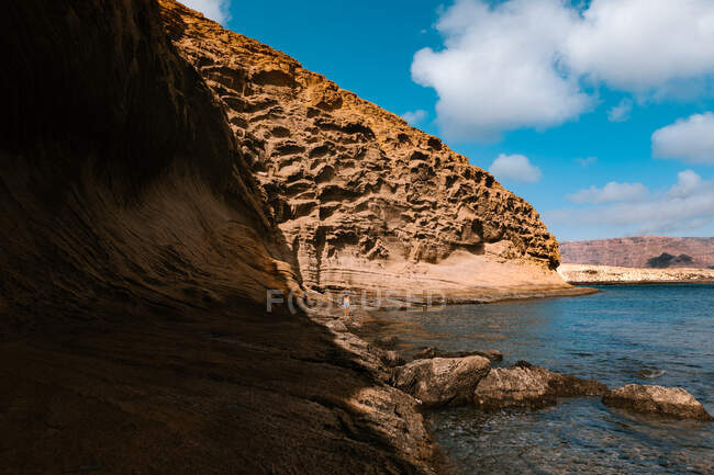 Female traveler standing on seashore with rocky cliffs near rippling sea during vacation in sunny exotic country — Stock Photo