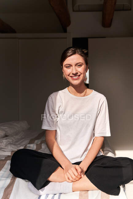 Full body of positive young lady in casual outfit and socks sitting on comfortable bed with crossed legs and smiling — Stock Photo