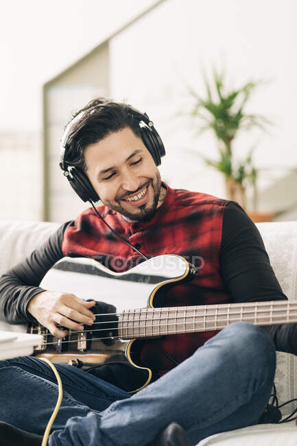 Content adult male artist in headset playing bass guitar on couch in house room in daylight — Stock Photo
