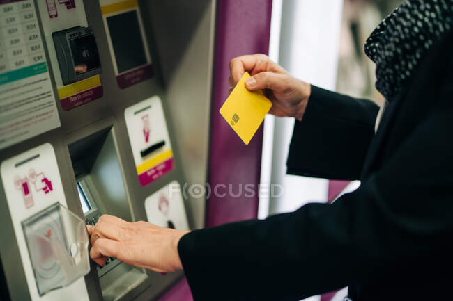 Vista lateral da fêmea cortada irreconhecível em pé perto da máquina ATM com cartão enquanto compra bilhete de trem enquanto está em pé no terminal moderno — Fotografia de Stock