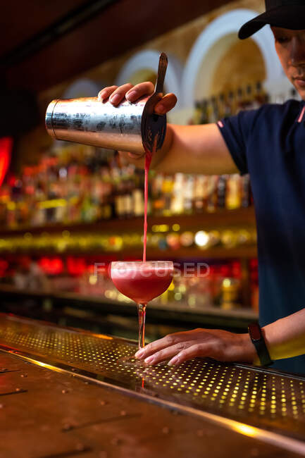 Joven camarero asiático trabajando en el bar con su agitador y vertiendo un cóctel en el vaso - foto de stock