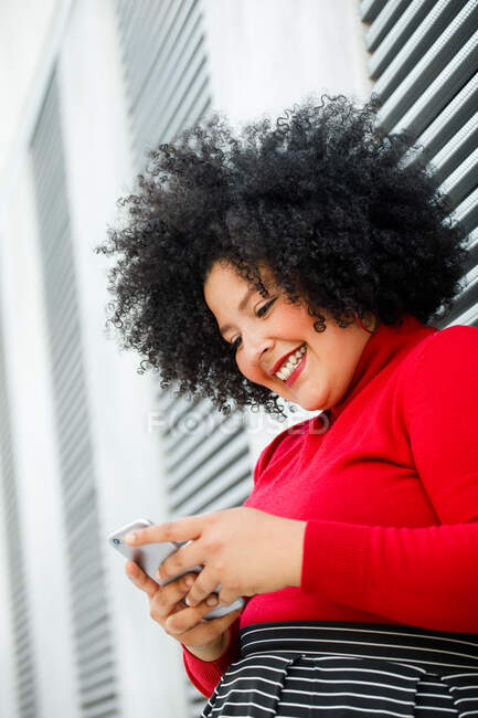 Вид збоку пухнастої усміхненої жінки в яскравому текстовому повідомленні на мобільний телефон, спираючись на ребристу стіну в місті — стокове фото