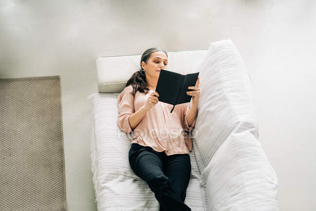 Зверху жінка читає книгу, лежачи на зручному дивані у вітальні з зеленою рослиною вдома — стокове фото