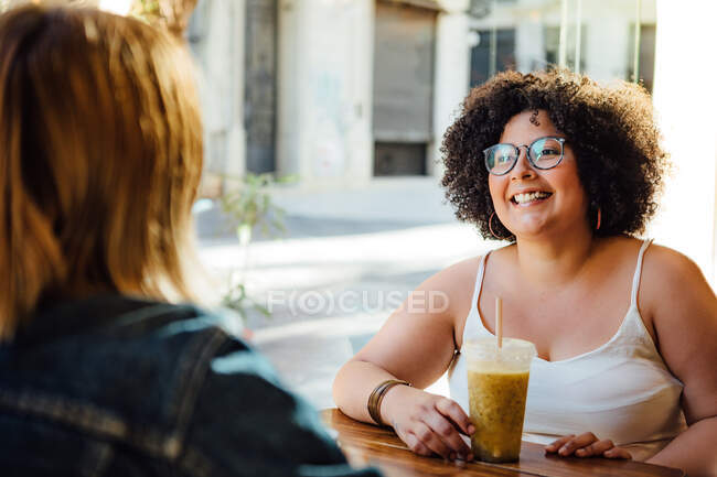 Искренняя женщина с освежающим напитком разговаривает с неузнаваемой партнершей за столом в уличной столовой — стоковое фото