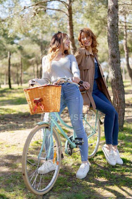 Full body of happy young girlfriends smiling and looking at each other on bicycle in sunny park in summer — Stock Photo
