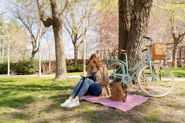 Full body of concentrated young female taking notes in notebook on checkered fabric with backpack near bicycle in park — Stock Photo