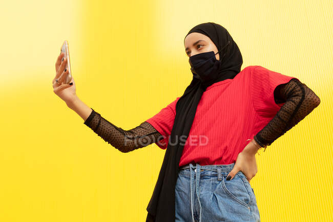 From below of cool ethnic female in headscarf with hand on hip taking self portrait on cellphone on yellow background — Stock Photo