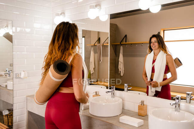 Mujer tranquila en ropa deportiva de pie con esterilla de yoga y toalla de sudor en el baño después del entrenamiento - foto de stock