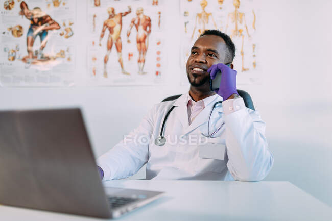 Alegre joven afroamericano médico en ropa médica y guantes teniendo conversación telefónica y sonriendo mientras está sentado en la mesa con el ordenador portátil y la taza de café durante el descanso en la clínica - foto de stock