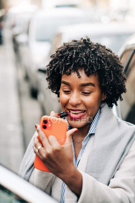 High angle of smiling African American female with curly hairstyle rouging lips with lipstick standing near car in traffic jam — Stock Photo