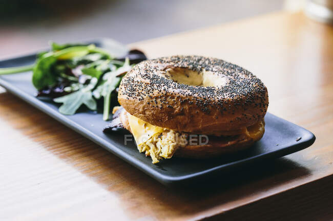 Appetizing bagel sandwich with cheese and chicken served on plate with rucola salad on table in cafe — Stock Photo