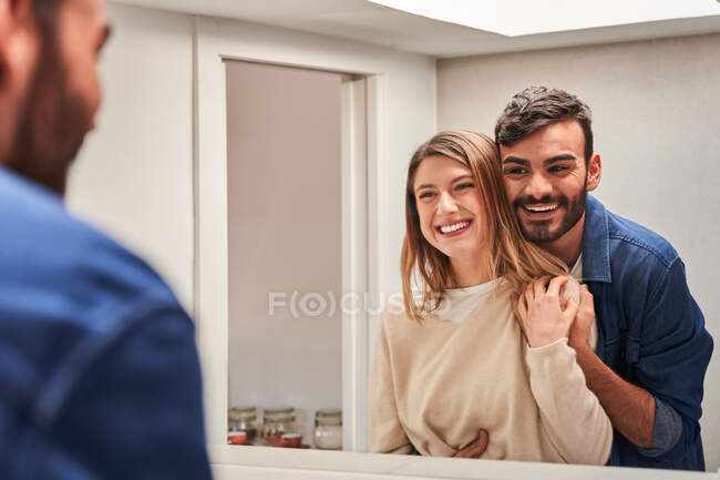 Cultiver joyeux jeune homme hispanique embrassant petite amie souriante tout en se tenant ensemble devant le miroir — Photo de stock