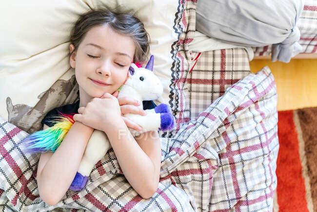 Vista dall'alto adorabile bambina che dorme tranquillamente su un letto accogliente e abbraccia morbido giocattolo unicorno in camera da letto leggera — Foto stock