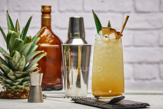 Cocktail giallo in vetro guarnito con pezzetto di ananas e foglie verdi con paglia di carta posta su sottobicchiere di ardesia con cucchiaio da bar — Foto stock