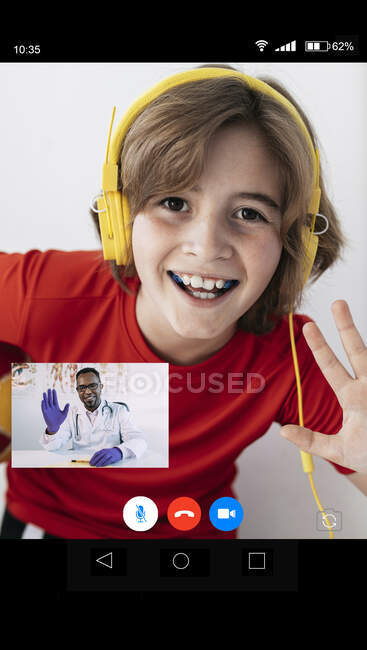 Cheerful boy in casual clothes and headphones waving hand and smiling while talking with African American doctor during online consultation — Stock Photo