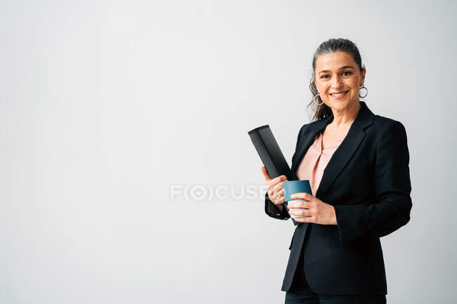 Позитивна середнього віку жінка-підприємець з хвостом у формальному костюмі, дивлячись на камеру, стоячи з папкою та чашкою напою на білому тлі — стокове фото