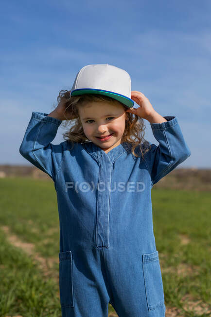 Happy girl in stylish clothes and cap looking at camera while standing on grass on sunny summer day in field — Stock Photo