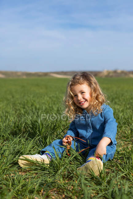 Мила щаслива дитина сидить у зеленому полі в сонячний день, дивлячись і граючи з травою влітку — стокове фото