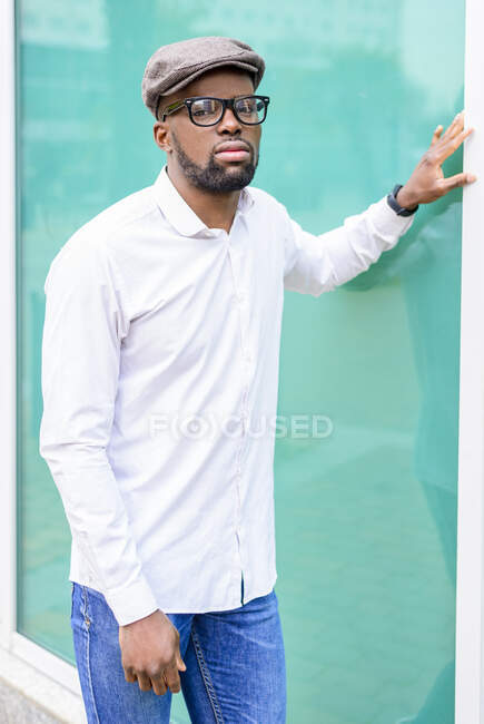 Autosicuro maschio afroamericano in abiti eleganti in piedi vicino a un moderno edificio di vetro sulla strada e guardando la fotocamera — Foto stock