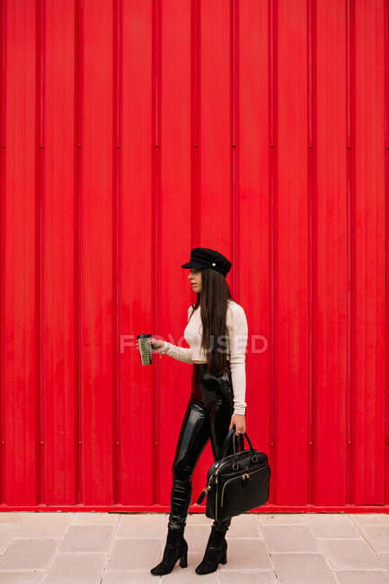Mujer de negocios segura en ropa de moda de pie con bebida para ir en la ciudad en el fondo de la pared roja y mirando hacia otro lado - foto de stock