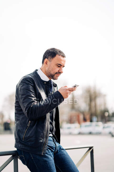 Seitenansicht der ethnischen männlichen Unternehmer mittleren Alters SMS auf dem Handy, während sie auf dem Zaun in der Stadt sitzen — Stockfoto