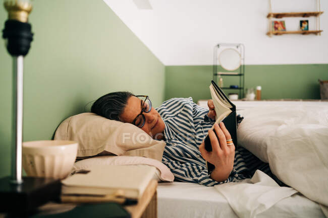 Femme hispanique mature détendue en vêtements décontractés et lunettes couchées au lit et lisant un livre intéressant avant de dormir — Photo de stock