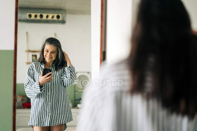 Cheerful middle aged female wearing striped blouse reflecting in mirror while taking self portrait on smartphone in room at home — Stock Photo