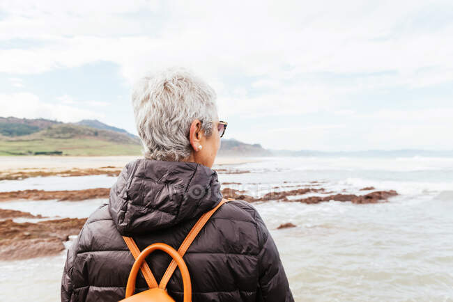 Back view of unrecognizable elderly female trekker in sunglasses with gray hair looking away against stormy ocean — Stock Photo