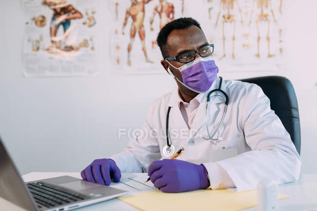 Médico afro-americano em óculos trabalhando com paciente on-line no netbook enquanto escrevia no arquivo do paciente à mesa no hospital — Fotografia de Stock