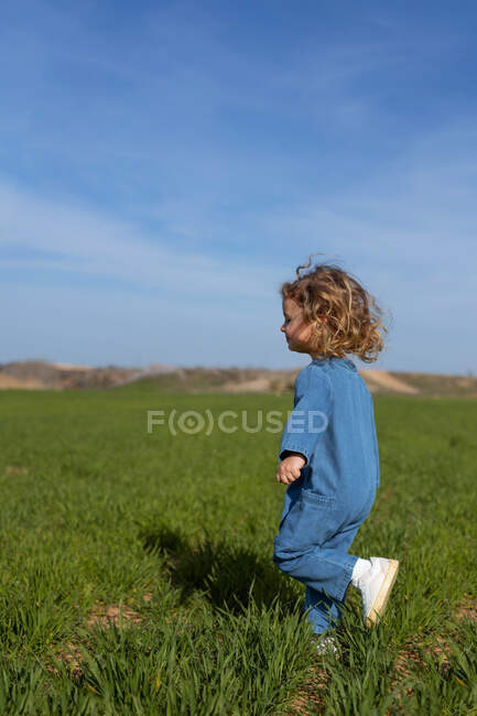 Вид сбоку восхищенной девушки с кудрявыми волосами, улыбающейся и бегущей по зеленой траве на фоне голубого неба в летний день в поле — стоковое фото